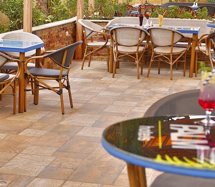 An outdoor patio area with round tables and wicker chairs, surrounded by a brick wall and greenery.