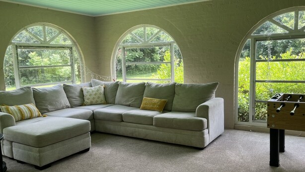 A cozy living room with a sectional sofa, a coffee table, and a foosball table. Large arched windows provide natural light and views of greenery outside.
