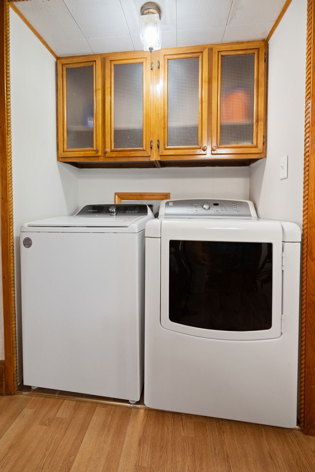 Une buanderie équipée de deux lave-linge et deux sèche-linge à chargement frontal, tous deux de couleur blanche, avec des armoires en bois au-dessus et un luminaire au plafond.