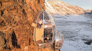 A transparent glass capsule attached to a cliffside, offering a view of a snowy landscape. - OVO Patagonia (El Chalten)