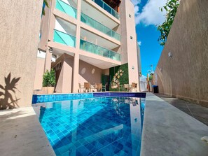 En moderne bygning med swimmingpool, udendørs siddepladser og en klar blå himmel.