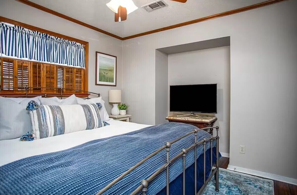 A bedroom with a bed, a TV, a window with blinds, a lamp, and a framed picture on the wall.