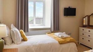 Une chambre d’hôtel comprenant un lit, une table de chevet, une lampe, une commode, un miroir, une fenêtre avec des rideaux et un téléviseur à écran plat.