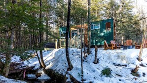 Eine Holzhütte im Wald mit einem kleinen Bach, einer Holzterrasse und Stühlen.