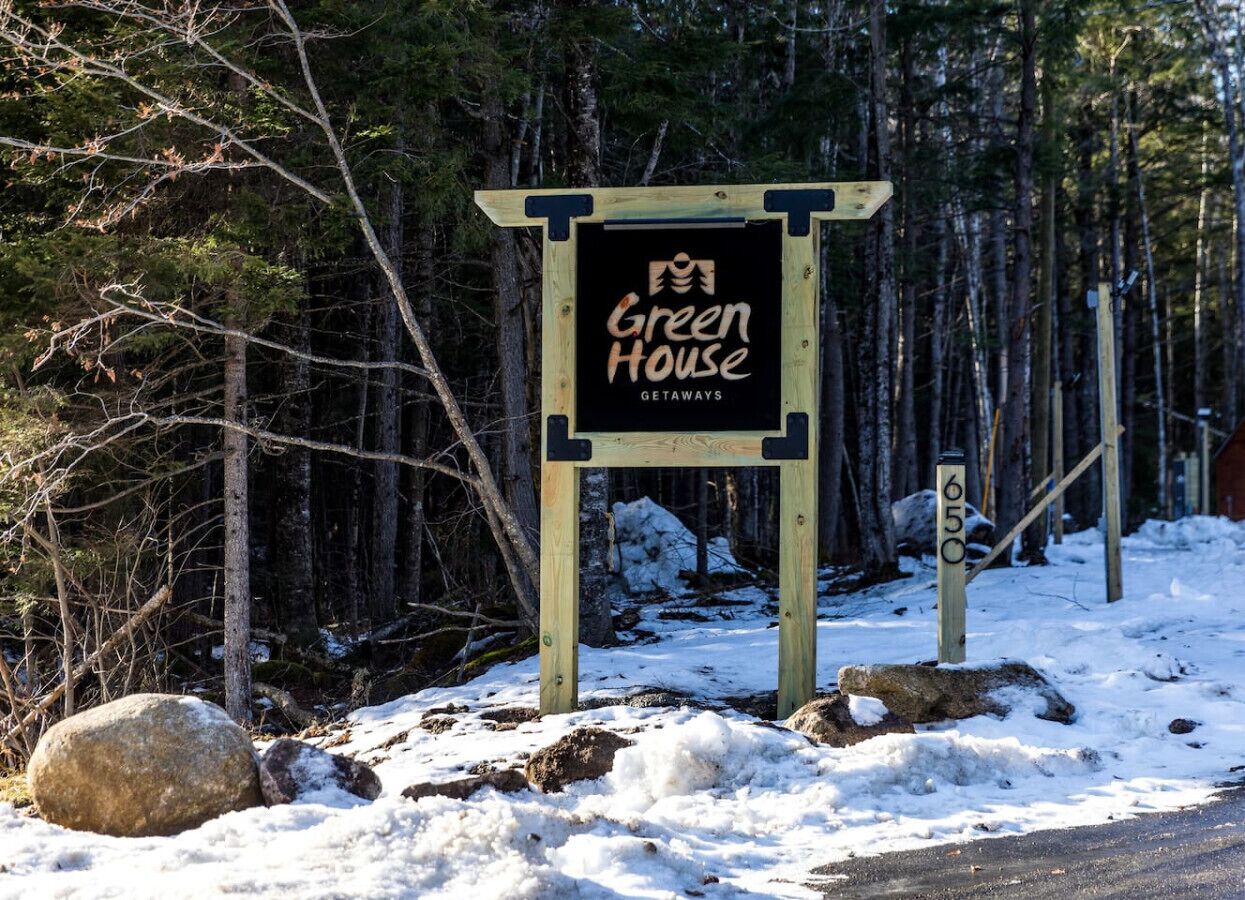 Un panneau en bois portant l’inscription « Green House Getaways » dans une zone boisée, la neige recouvrant le sol.
