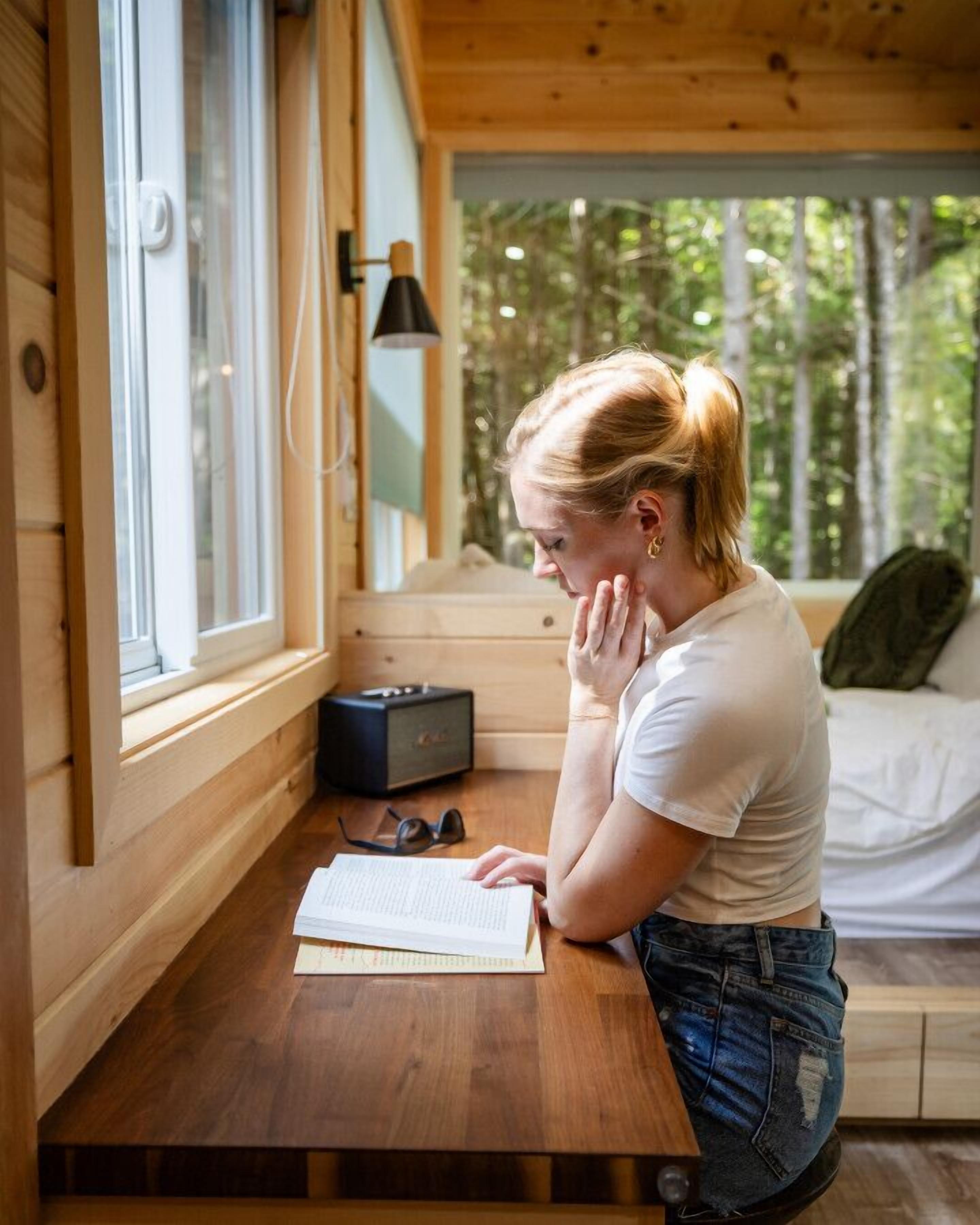 Una persona asseguda a un escriptori de fusta en una cabana, amb una finestra, una làmpada i un llit al fons.