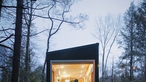 Una cabaña pequeña y moderna con un frente de vidrio, ubicada en un bosque nevado.