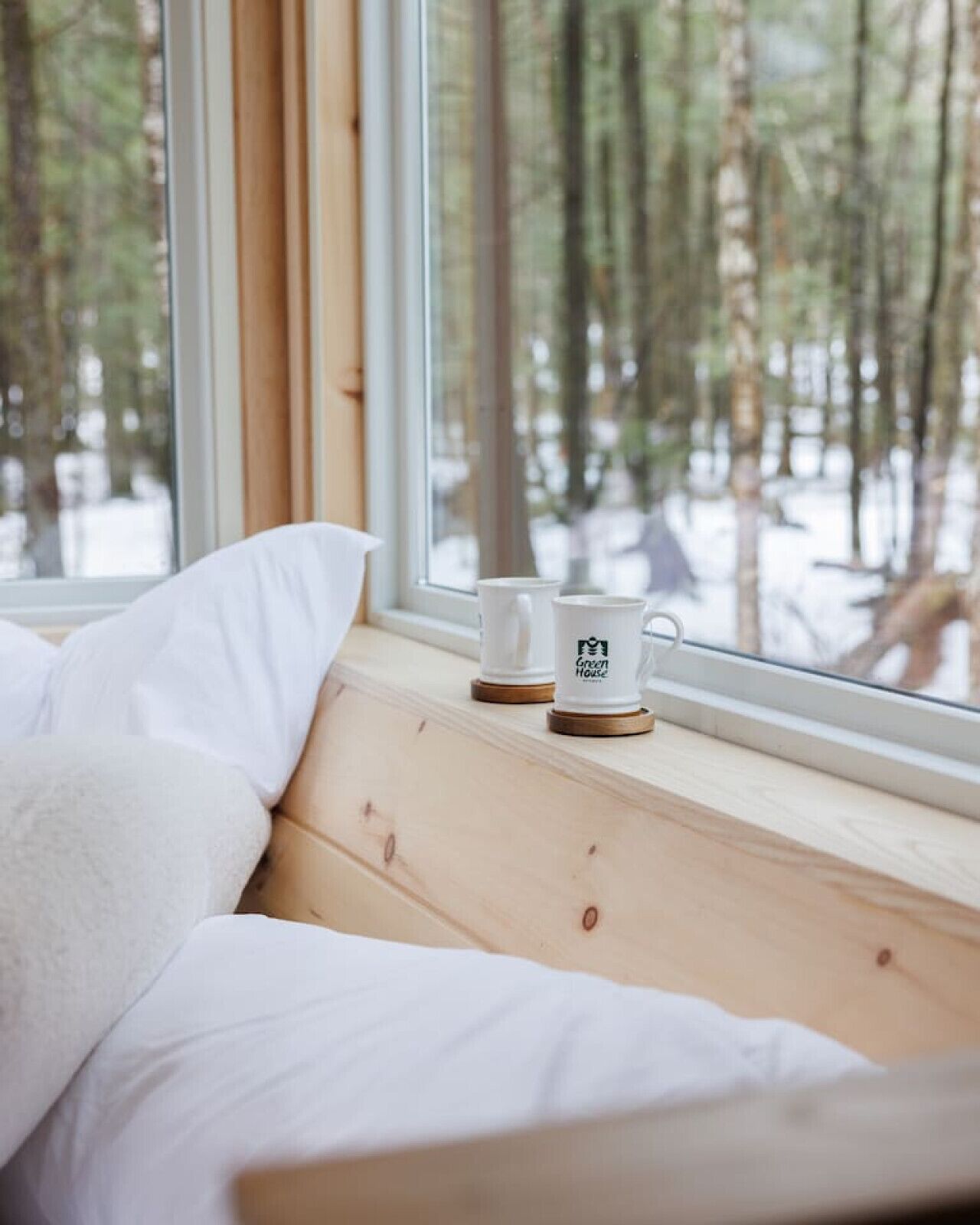 A bed with white bedding, a window with a view of a forest, and two white mugs on the windowsill.