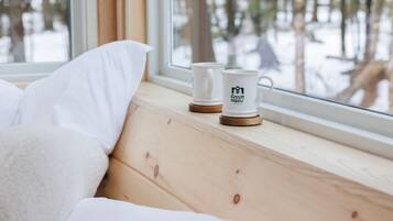 A bed with white bedding, a window with a view of a forest, and two white mugs on the windowsill.