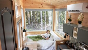 Una cabaña pequeña con cama, un escritorio, un lavamanos y ventana con vistas a un bosque.