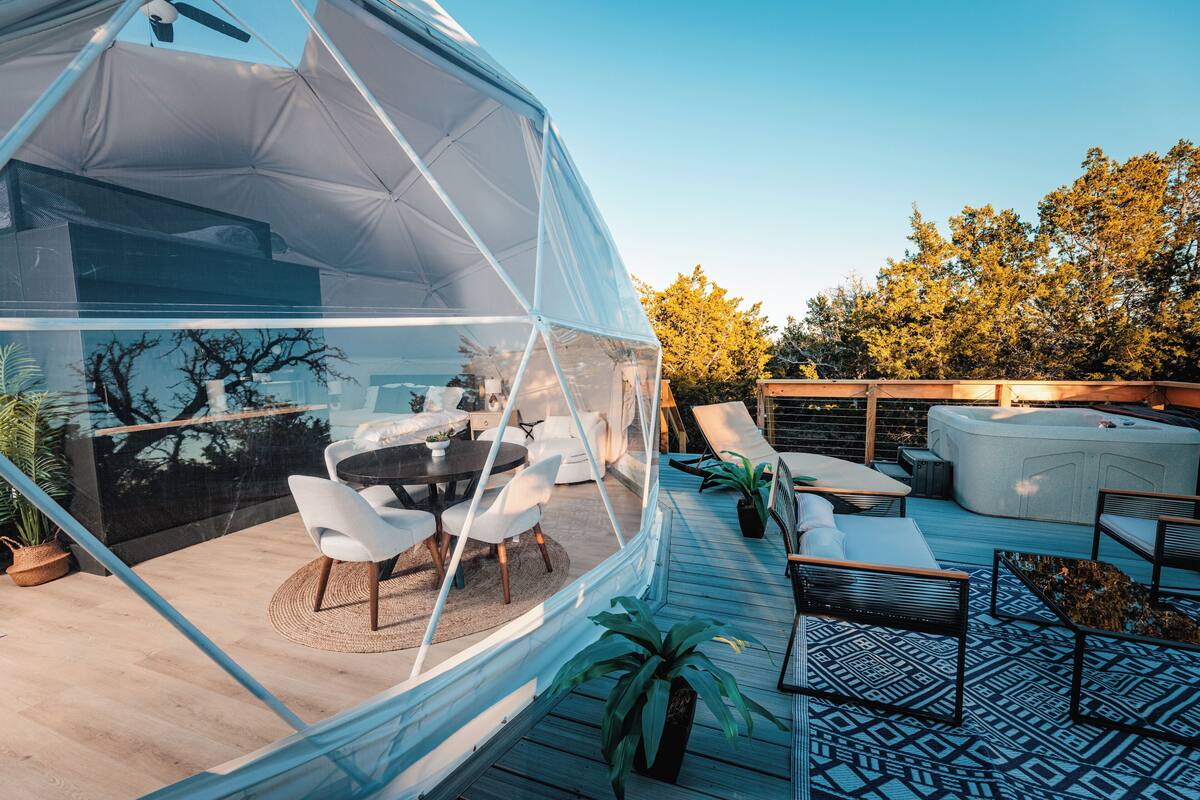 A modern outdoor patio with a glass enclosure, white furniture, and a wooden deck.