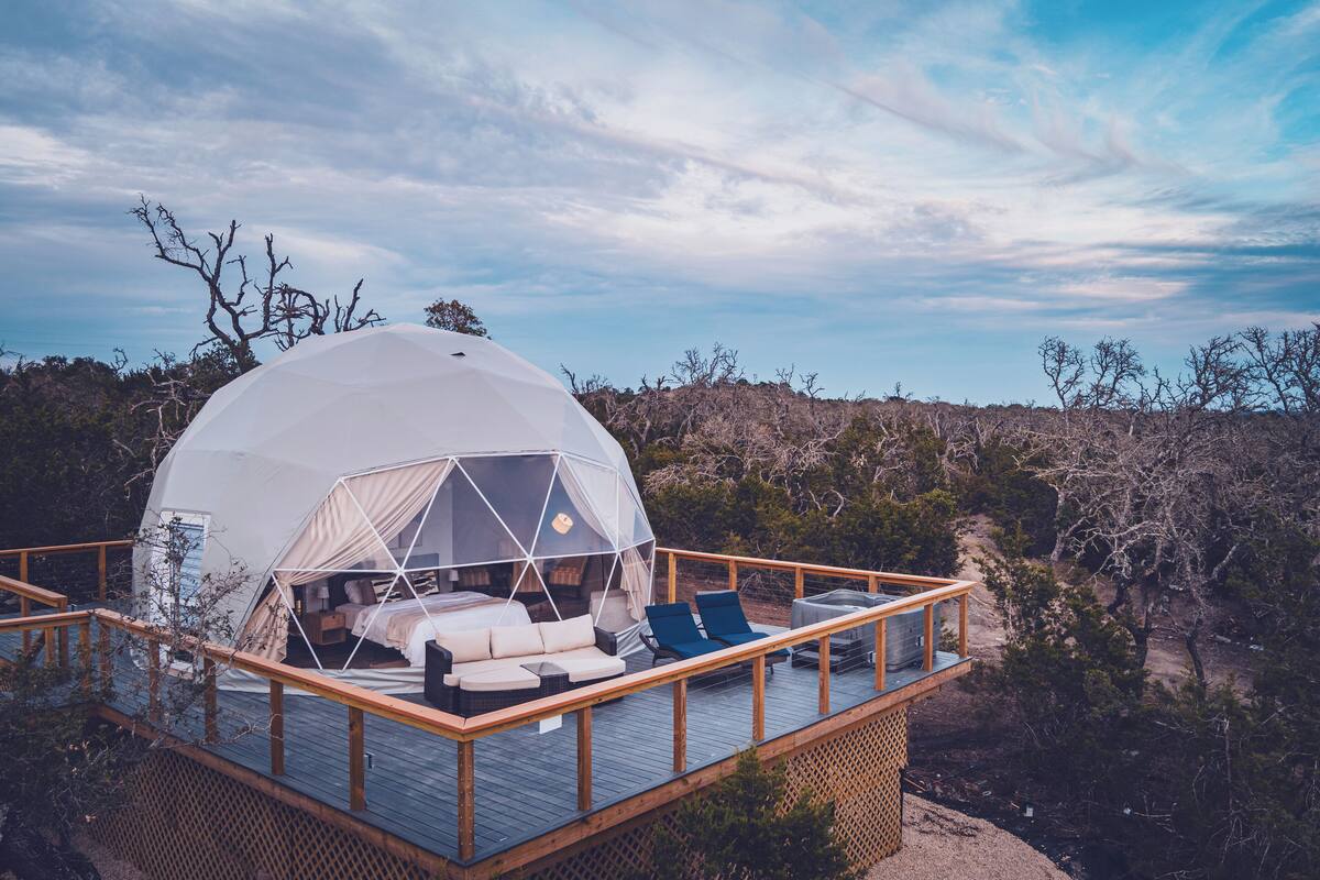 A dome-shaped tent with a bed and seating area on a wooden deck, surrounded by trees.