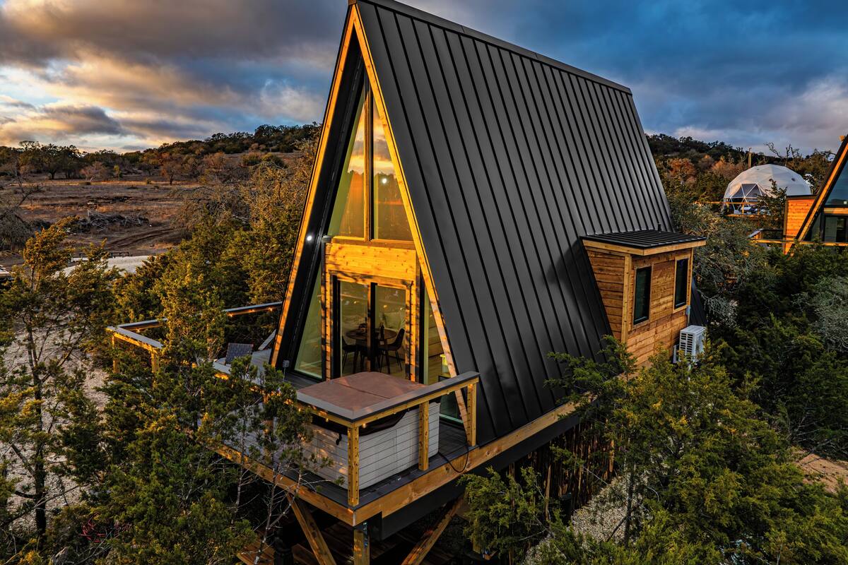 A modern, triangular-shaped house with a large glass window and a deck, surrounded by trees and a dome structure in the background.