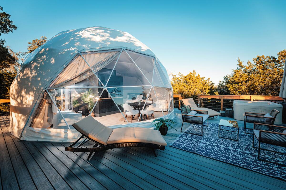 A geodesic dome structure with a transparent roof, furnished with lounge chairs, a small table, and a potted plant.