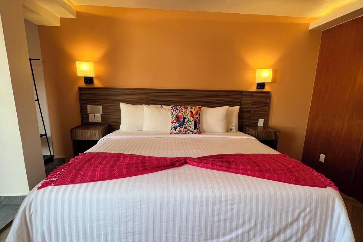 Habitación de hotel con una cama grande, dos lámparas de noche, un cabecero de madera y una almohada decorativa roja.