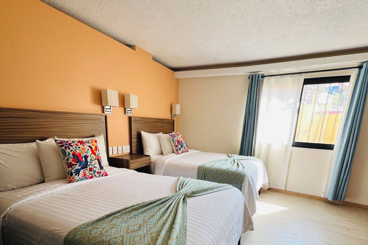 Habitación de hotel con dos camas, cabecero de madera, mesita de noche, lámparas de pared y una ventana con cortinas.