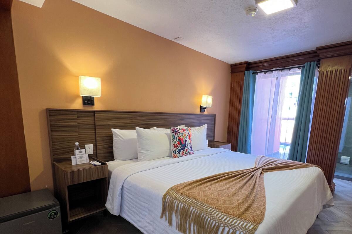 Habitación de hotel con una cama grande, mesitas de noche, lámpara, refrigerador y una ventana con cortinas.
