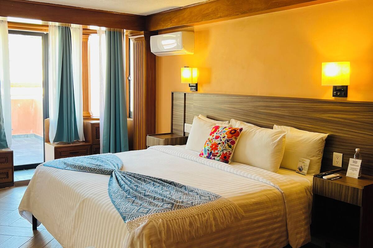 Habitación de hotel con una cama grande, mesitas de noche, un equipo de aire acondicionado y vistas al exterior.