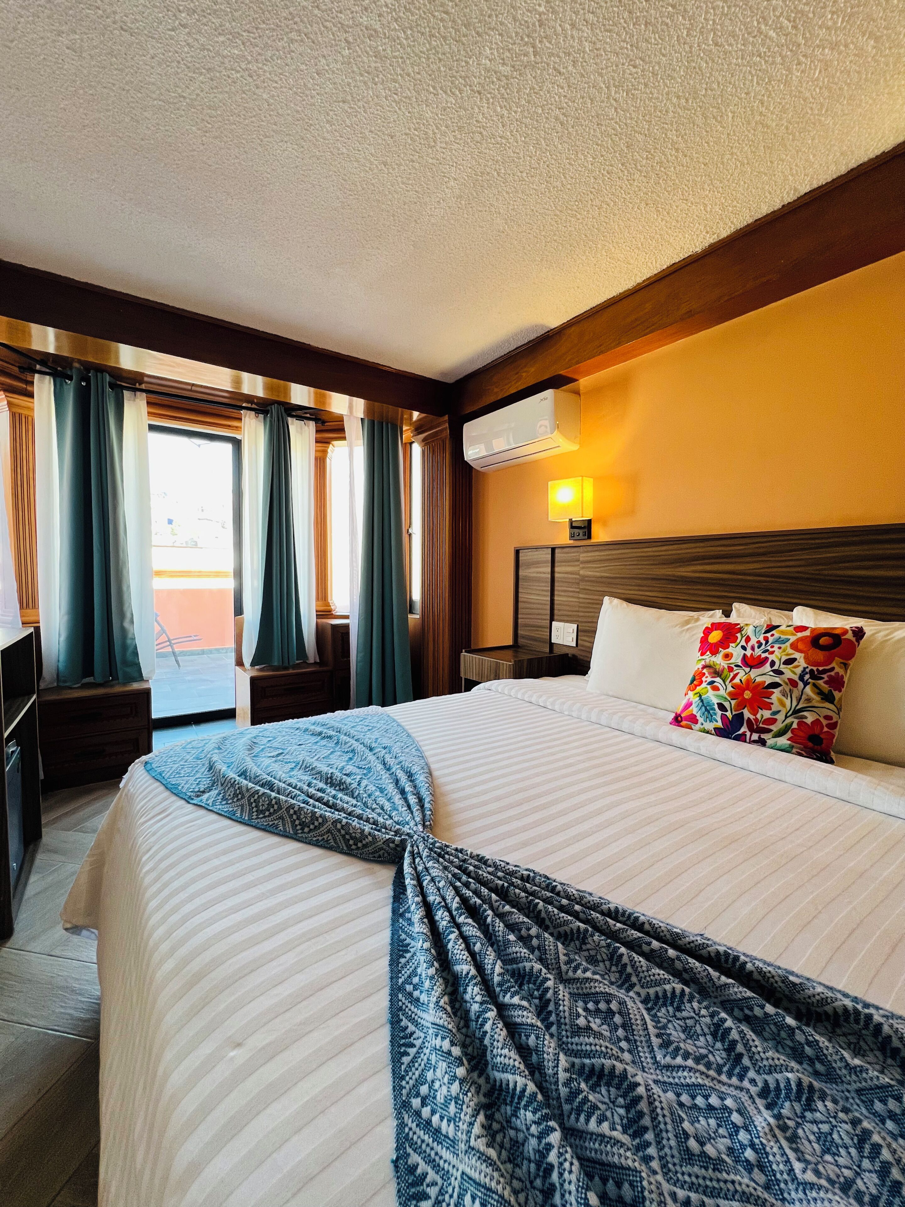 A hotel room with a large bed, a bedside table, a lamp, and a view of the outside.