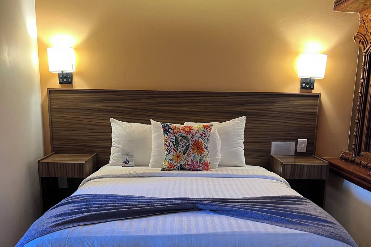 Habitación de hotel con cama, dos mesitas de noche, un espejo y dos lámparas de pared.