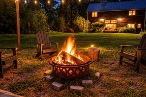 A fire pit with burning logs, surrounded by wooden chairs, set on a grassy lawn with a house in the background. - Hudson Retreat - 1780s Renovated Barn (Hudson)