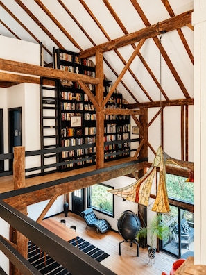 A spacious loft with exposed wooden beams, a large bookshelf, a hanging light fixture, and a view of the outdoors. - Hudson Retreat - 1780s Renovated Barn (Hudson)