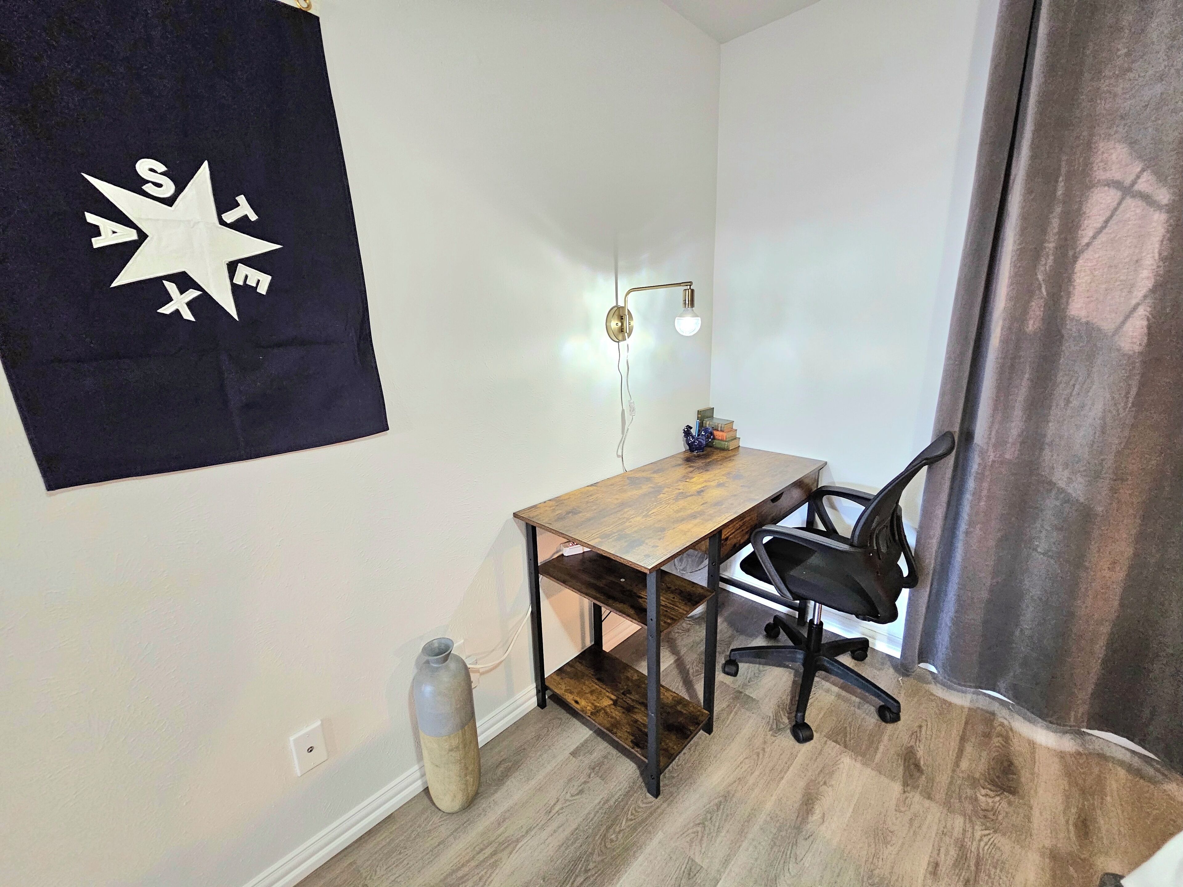 Um pequeno espaço de escritório com uma mesa de madeira, uma cadeira de escritório preta, uma luminária na parede e um vaso decorativo.