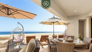 Un patio frente a la playa con muebles de mimbre, una silla colgante y sombrillas que dan sombra.