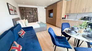 Una sala de estar moderna con un sofá azul, una mesa de comedor con sillas, una zona de cocina con armarios de madera y un televisor al fondo.