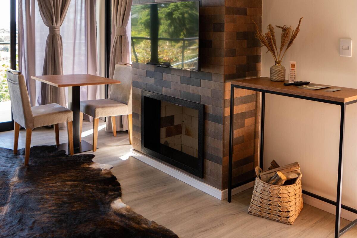 A modern living room with a brick fireplace, a flat-screen TV, a dining table with chairs, and a large rug.