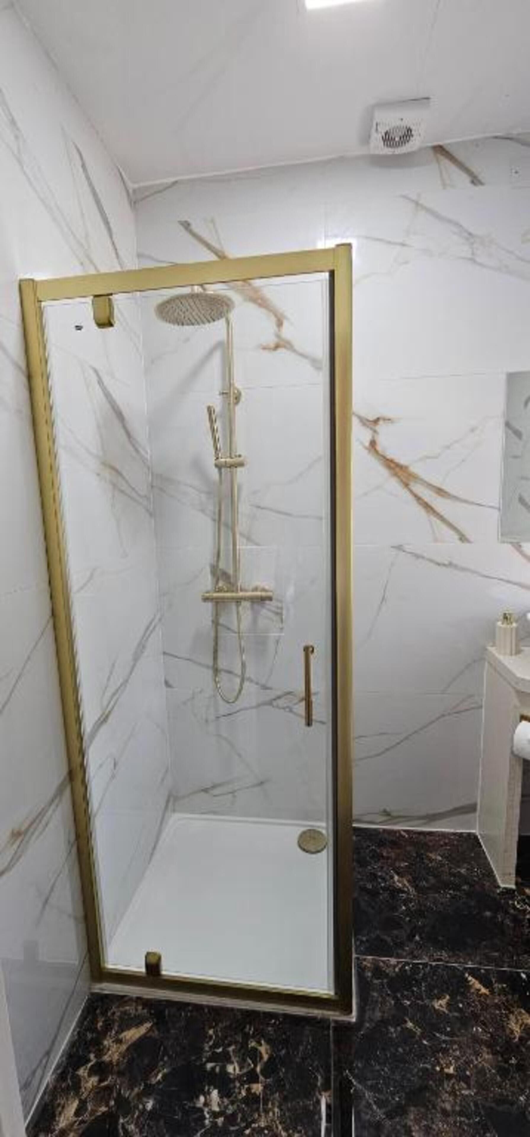 Una ducha con cerramiento de vidrio, marco dorado, paredes de mármol y suelo de mármol negro.