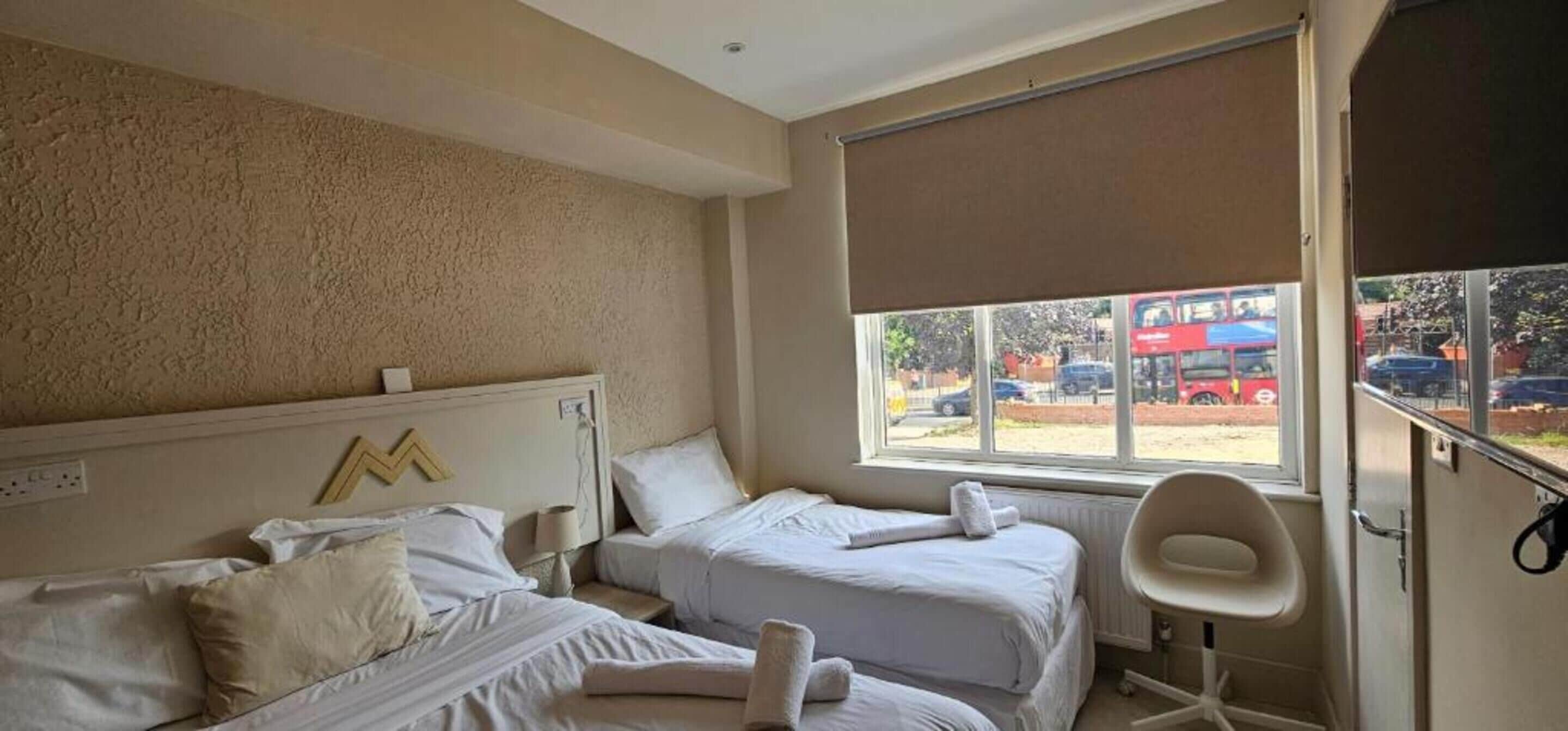 Una camera d'albergo con due letti, una finestra che mostra un autobus rosso e auto, e una sedia bianca.