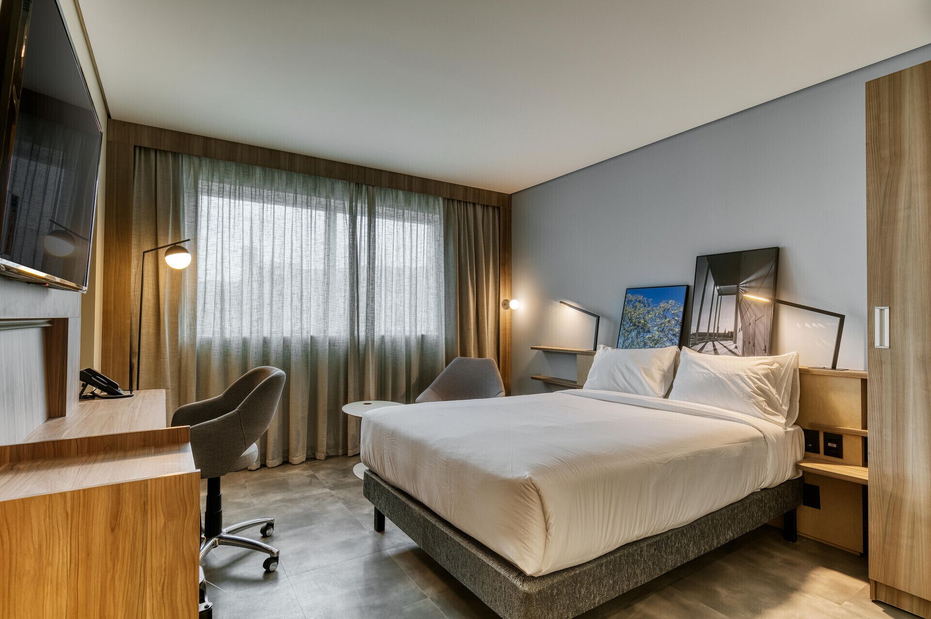 Habitación de hotel con cama, escritorio, silla, mesitas de noche y un ventanal con cortinas.