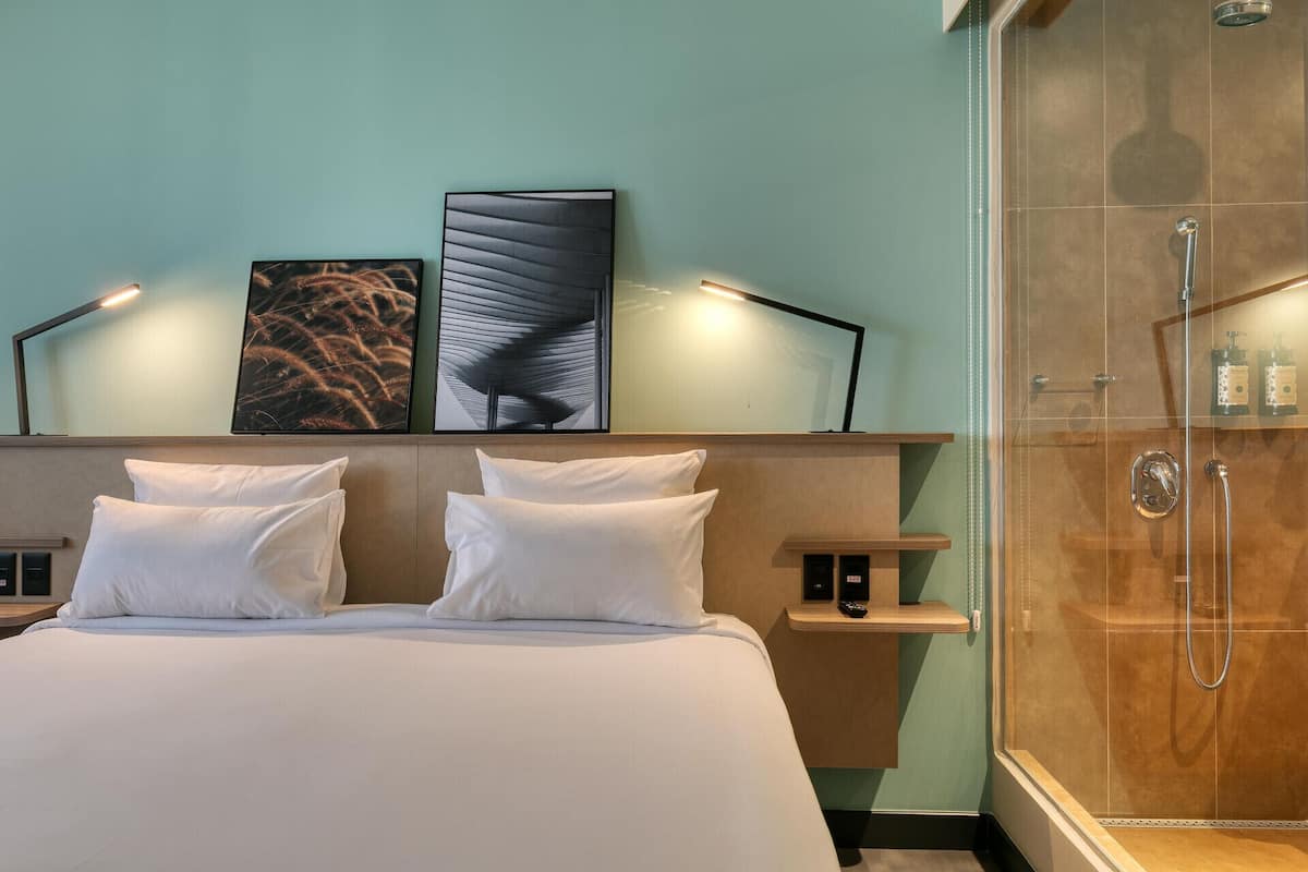 Une chambre d’hôtel comprenant un lit, deux tables de chevet, un grand miroir et une douche séparée par des parois en verre.