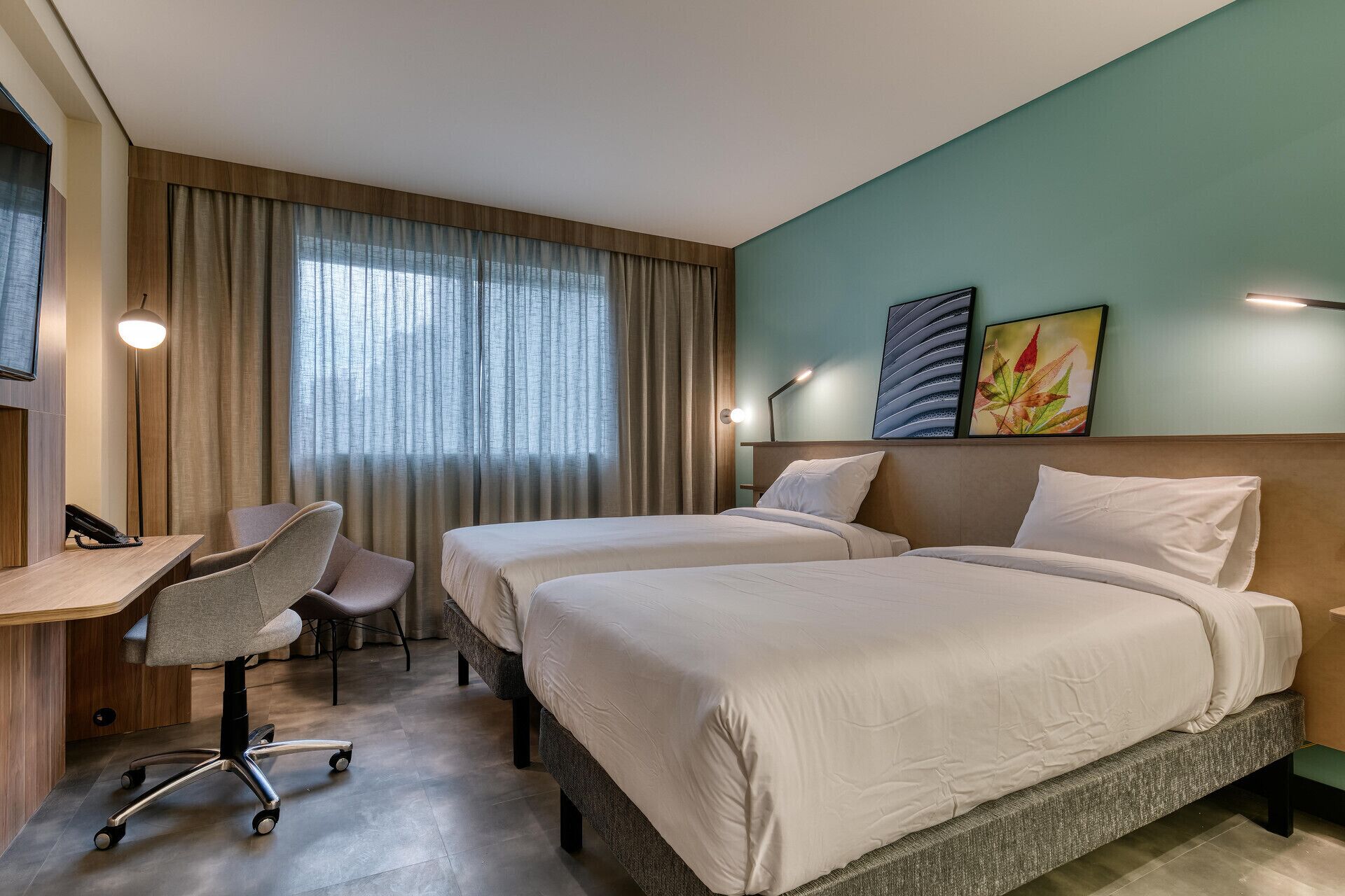 Quarto de hotel com duas camas, uma escrivaninha, uma cadeira e uma janela grande com cortinas.