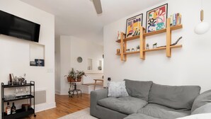 A modern living room with a grey sofa, a black TV on the wall, wooden shelves with books and plants, and a small table with a plant. - Best Denver Neighborhood! Walkable to EVERYTHING!  (Denver)