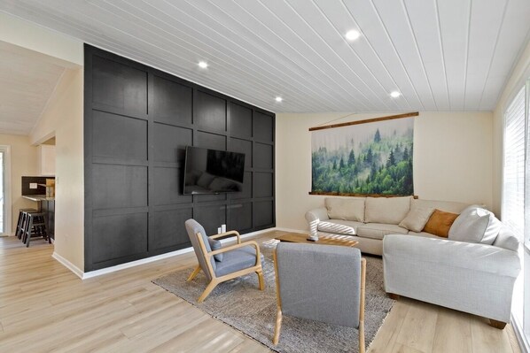 Un salon moderne avec un téléviseur à écran plat fixé sur un mur recouvert de panneaux sombres, un canapé gris, un divan de couleur claire et un plancher en bois.