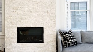 A living room with a stone wall, a fireplace, a grey sofa, and a window. - Charming Cottage steps to restaurants, shops & slopes (Park City)