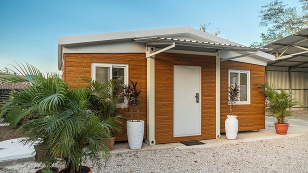 Une maison moderne de plain-pied, avec une façade en bois, une porte blanche et deux fenêtres. Des plantes en pot sont disposées devant la maison.