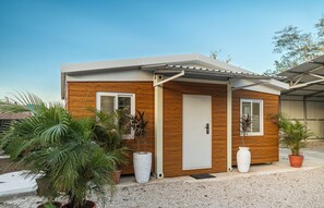 Une maison moderne de plain-pied, avec une façade en bois, une porte blanche et deux fenêtres. Des plantes en pot sont disposées devant la maison.