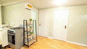Une cuisine compacte équipée d’une cuisinière à gaz, d’un four et d’une petite étagère avec des plantes. Les portes sont blanches et le sol est en bois.