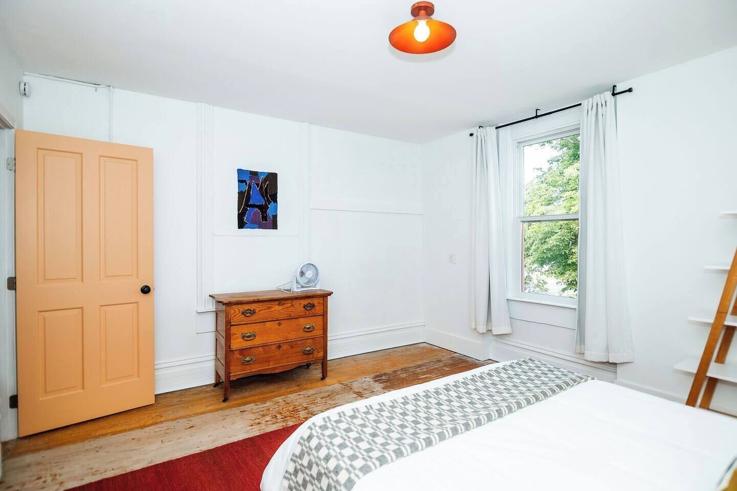 A bedroom with a wooden dresser, a bed with a patterned bedspread, a window with curtains, and a ceiling light.