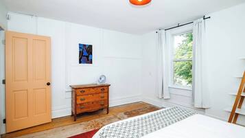 A bedroom with a wooden dresser, a bed with a patterned bedspread, a window with curtains, and a ceiling light.