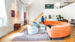 A spacious living room with a large orange sofa, a blue armchair, and a patterned rug. A staircase with a yellow railing leads to an upper level. - Find Your Sanctuary at The Church (Helena)