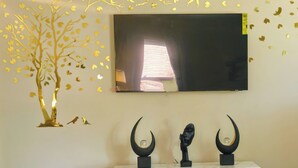 Un salon moderne doté d’un téléviseur à écran plat fixé au mur, d’une console blanche ornée de trois sculptures et d’un motif décoratif représentant un arbre aux feuilles dorées.