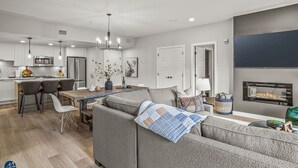 A modern kitchen with white cabinets, stainless steel appliances, and pendant lighting. There is a dining area with a wooden table and chairs, and a living room with a grey sofa and a fireplace.