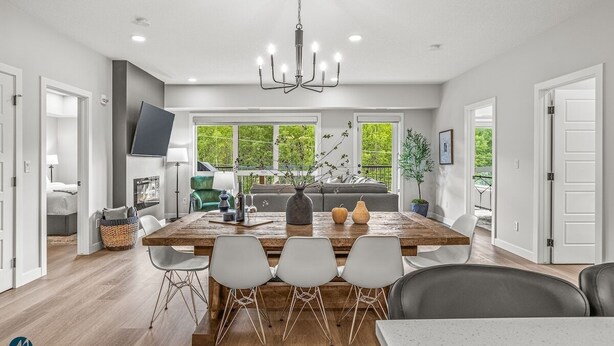 A modern kitchen with a large wooden table, white chairs, and a chandelier. There is a sofa and a TV in the background.