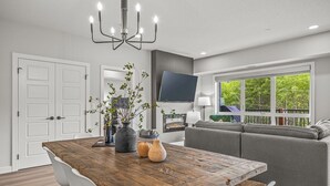 A modern living room with a wooden dining table, white chairs, a grey sofa, and a large window with a view of greenery.