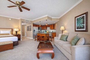 A hotel room with a bed, a sofa, a coffee table, a dining area with a kitchen in the background, and a ceiling fan.