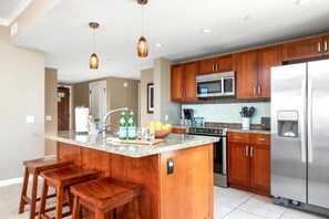 Eine Küche mit Holzausstattung, einem Edelstahlkühlschrank, einer Mikrowelle und einer Arbeitsplatte aus Granit. Zwei Barhocker und ein Spülbecken mit Armatur sind ebenfalls vorhanden.